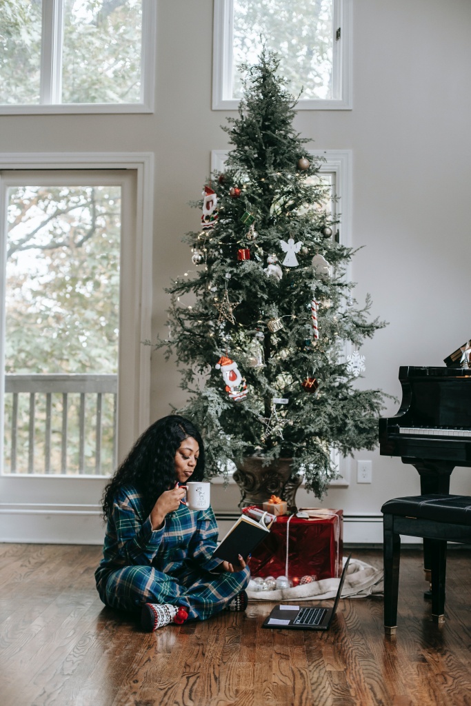 A woman reading a book and sipping her coffee near a christmas tree