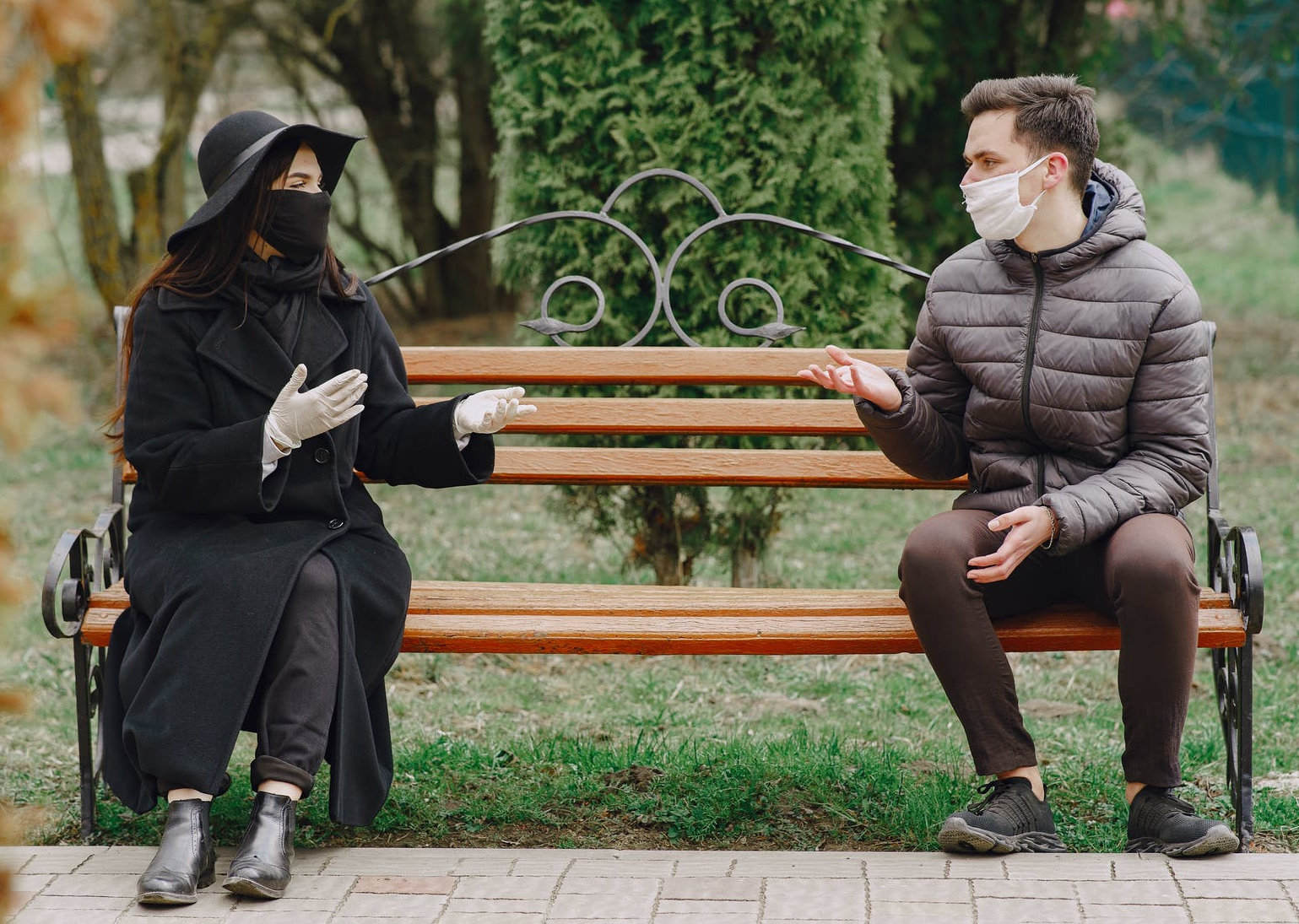 Two people sitting on bench wearing masks and social distancing